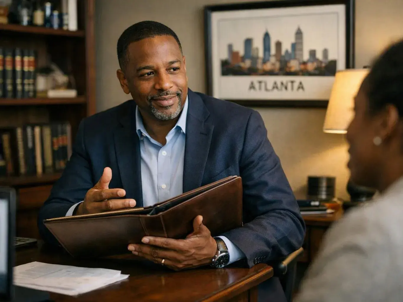 A man in a navy suit talks while holding a brown folder across a desk in a cozy office