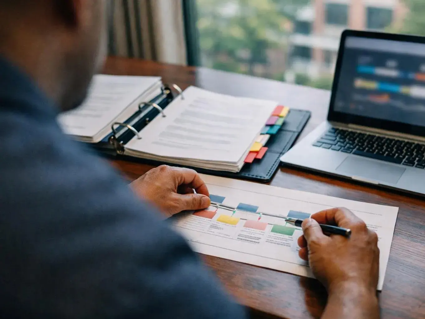 A person marks a printed chart beside a laptop and open binder at a wooden desk