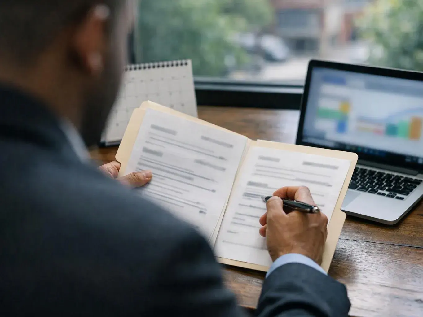 A person in a suit writes in a notebook beside a laptop at a wooden desk by a window